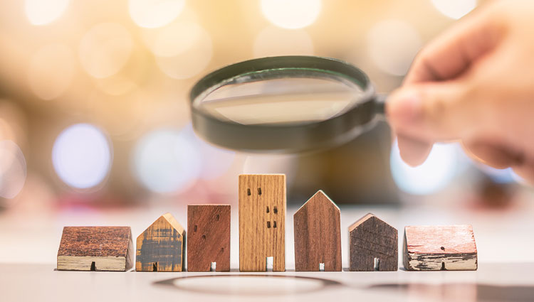 Hand holding a magnifying glass over miniature wooden houses, symbolizing estate planning, property evaluation, and identifying assets for bequest marketing.